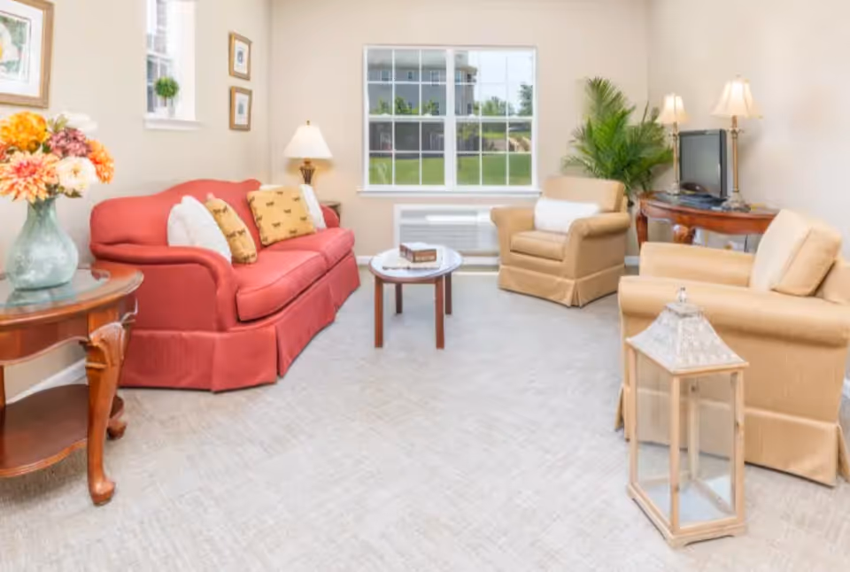 Bright living room with a red sofa, beige armchairs, coffee table, side tables and a window overlooking greenery.