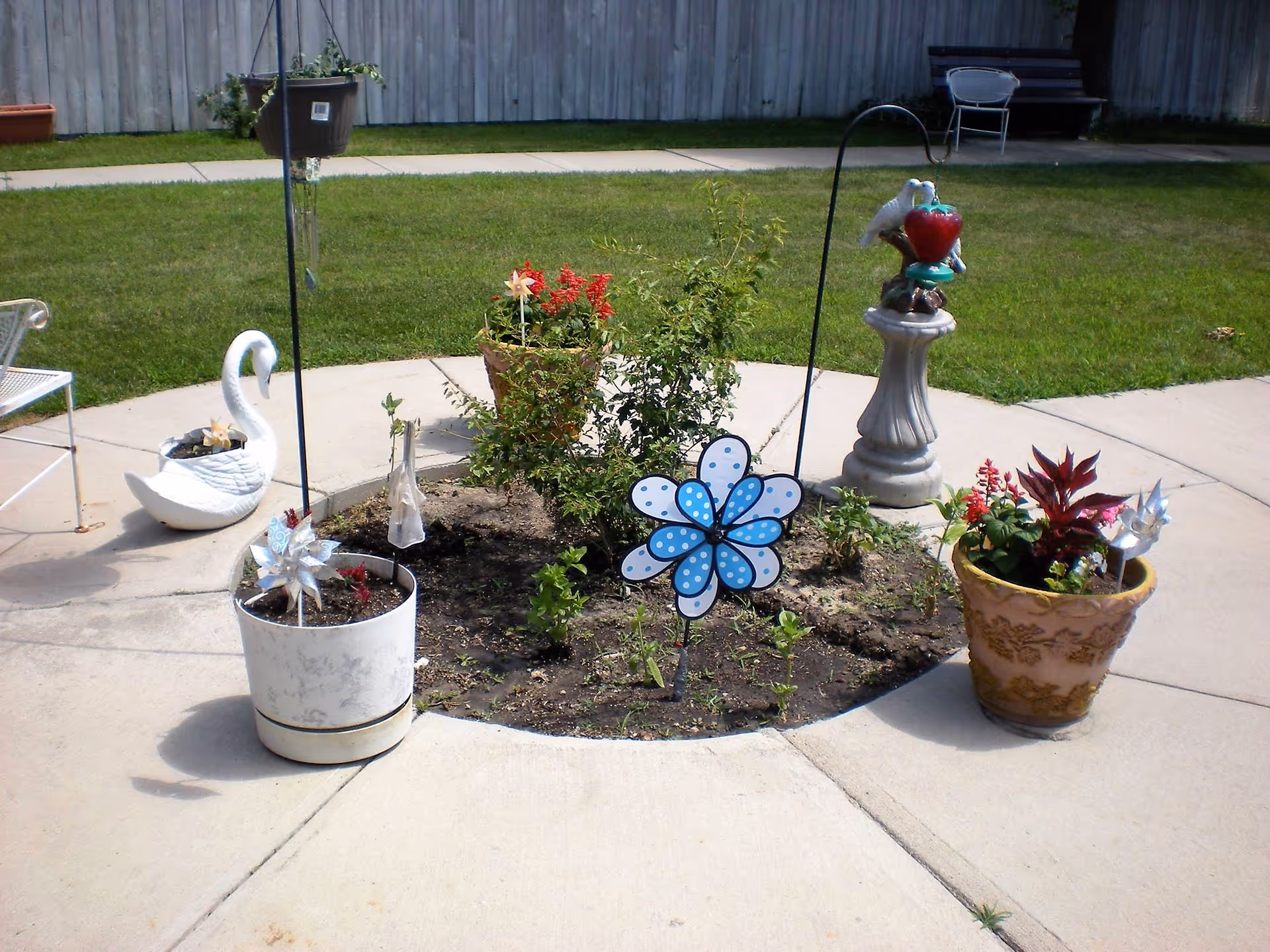 A small circular garden bed with various plants and flowers surrounded by a concrete walkway. Decorative items include a blue and white polka dot pinwheel, a white swan planter, a bird feeder on a pedestal, and several potted plants. In the background, there is a wooden fence, a bench, and a white chair on a grassy lawn.