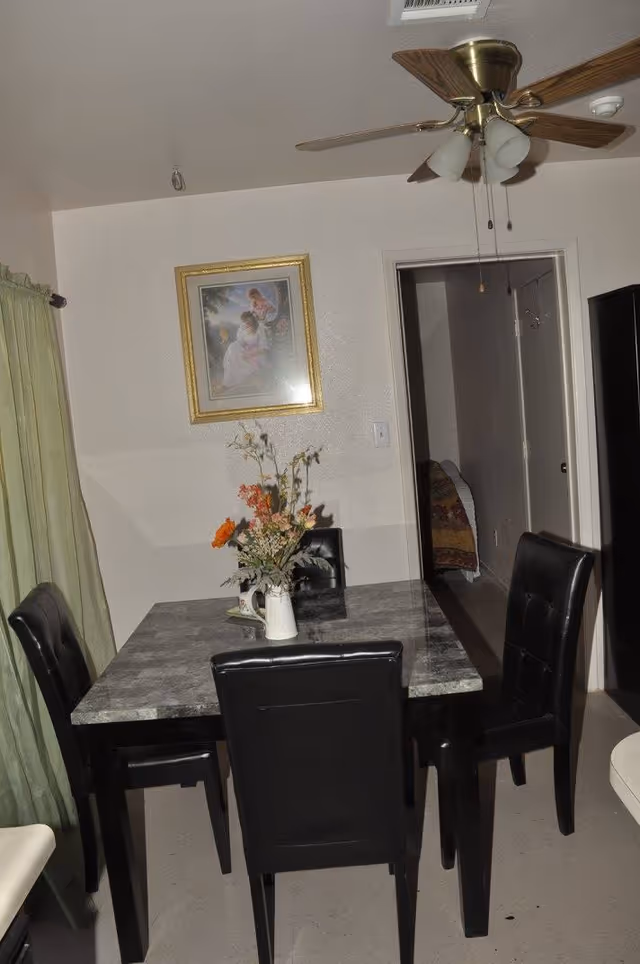 Small dining area with a square marble-top table, four black chairs, a vase of flowers, a ceiling fan, and a framed picture on the wall.