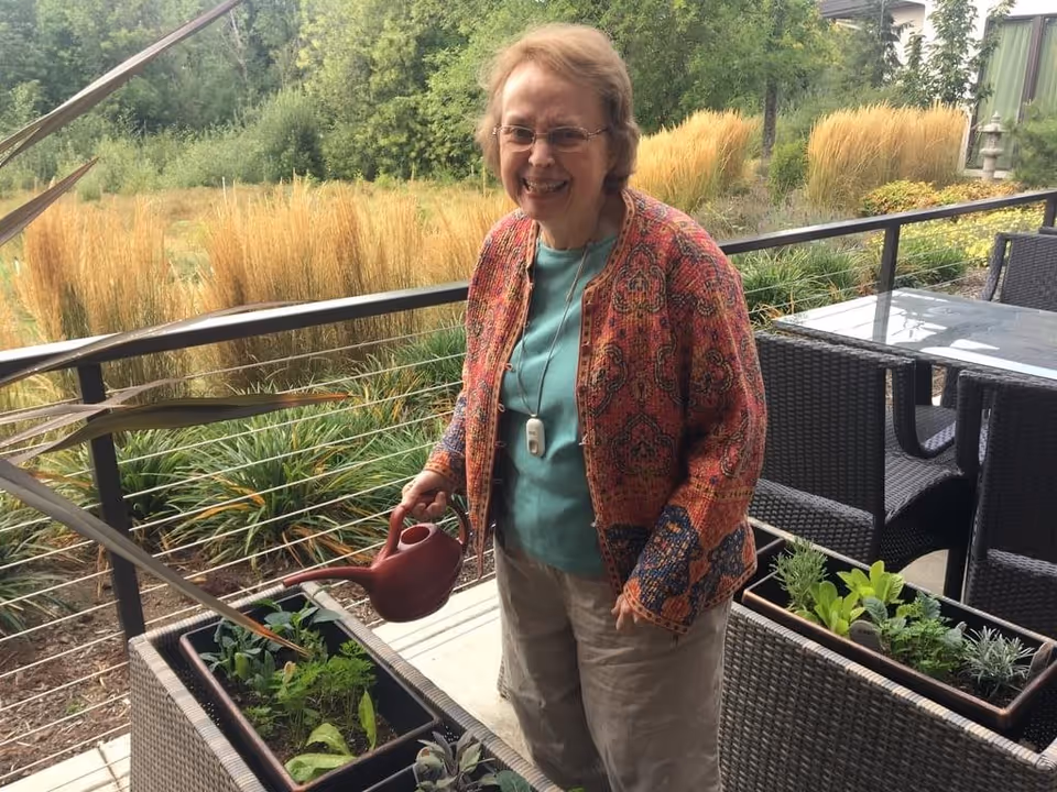 An elderly woman wearing glasses and a colorful patterned jacket is smiling while watering plants in rectangular planters on a patio. Behind her, there is outdoor furniture and a natural landscape with tall grasses and trees.