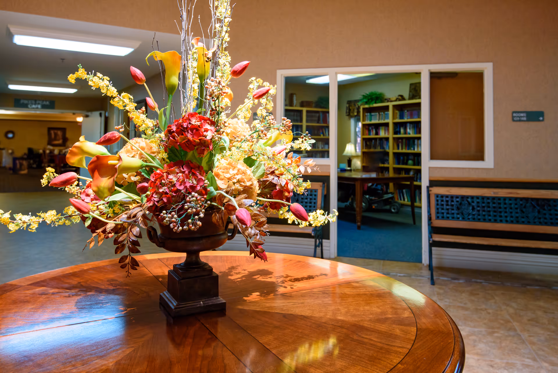 Large floral arrangement on a round wooden table in a nursing center foyer with a library room visible in the background.