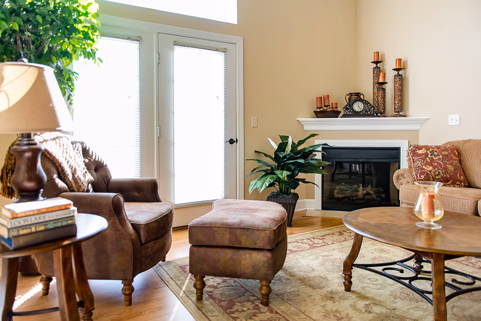 A cozy living room with a brown armchair and matching ottoman, a wooden side table with a lamp and stacked books, a beige sofa with a decorative pillow, a wooden coffee table with a glass candle holder, a fireplace with candles and a clock on the mantel, and a large green plant near glass doors with blinds.