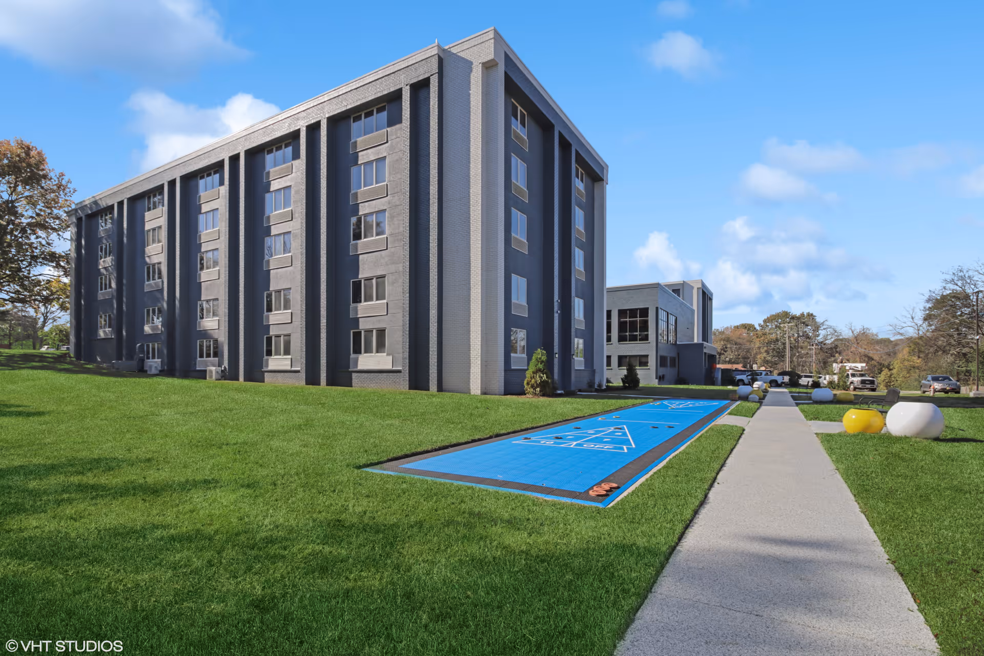 Exterior view of a multi-story senior living facility building with a well-maintained green lawn and a blue shuffleboard court. A concrete walkway runs alongside the lawn, leading towards the building entrance. The sky is clear with a few clouds.