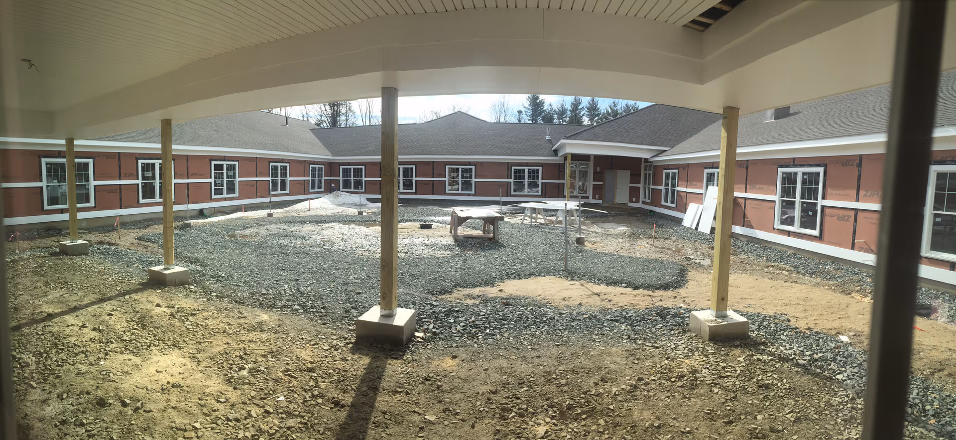 View of an unfinished courtyard area surrounded by a single-story building under construction, with gravel and dirt on the ground and wooden support posts holding up a covered walkway.