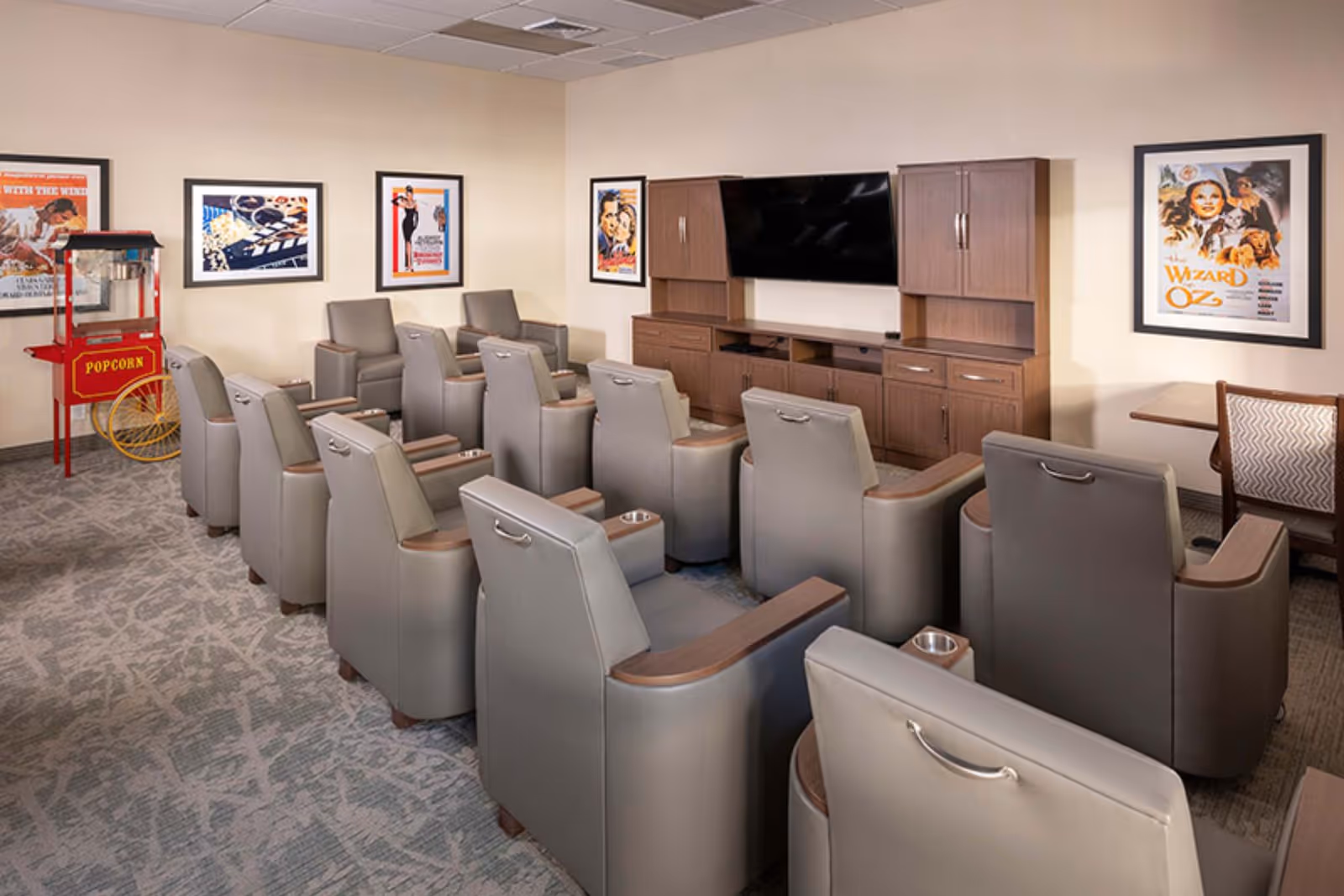 A small movie theater room with two rows of gray recliner chairs facing a flat-screen TV mounted on a wooden cabinet. The room has framed vintage movie posters on the walls and a red popcorn machine in the corner.