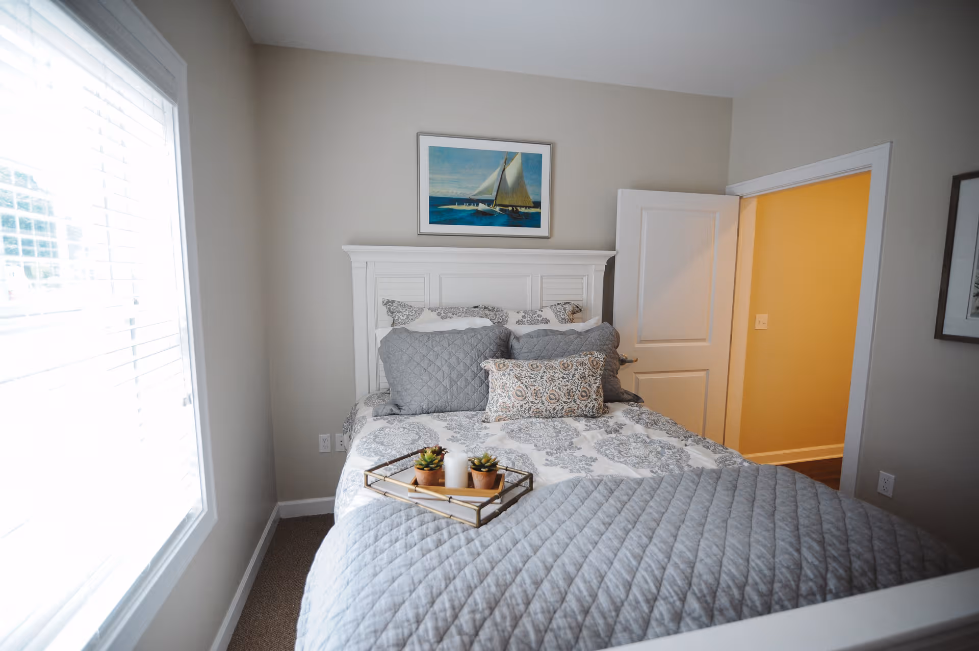 A cozy bedroom with a neatly made bed featuring a gray quilted blanket and patterned pillows. A tray with small potted plants and candles is placed on the bed. The room has light-colored walls, a white headboard, a window with blinds on the left, and an open door leading to a hallway with yellow walls. A framed picture of a sailboat hangs above the headboard.