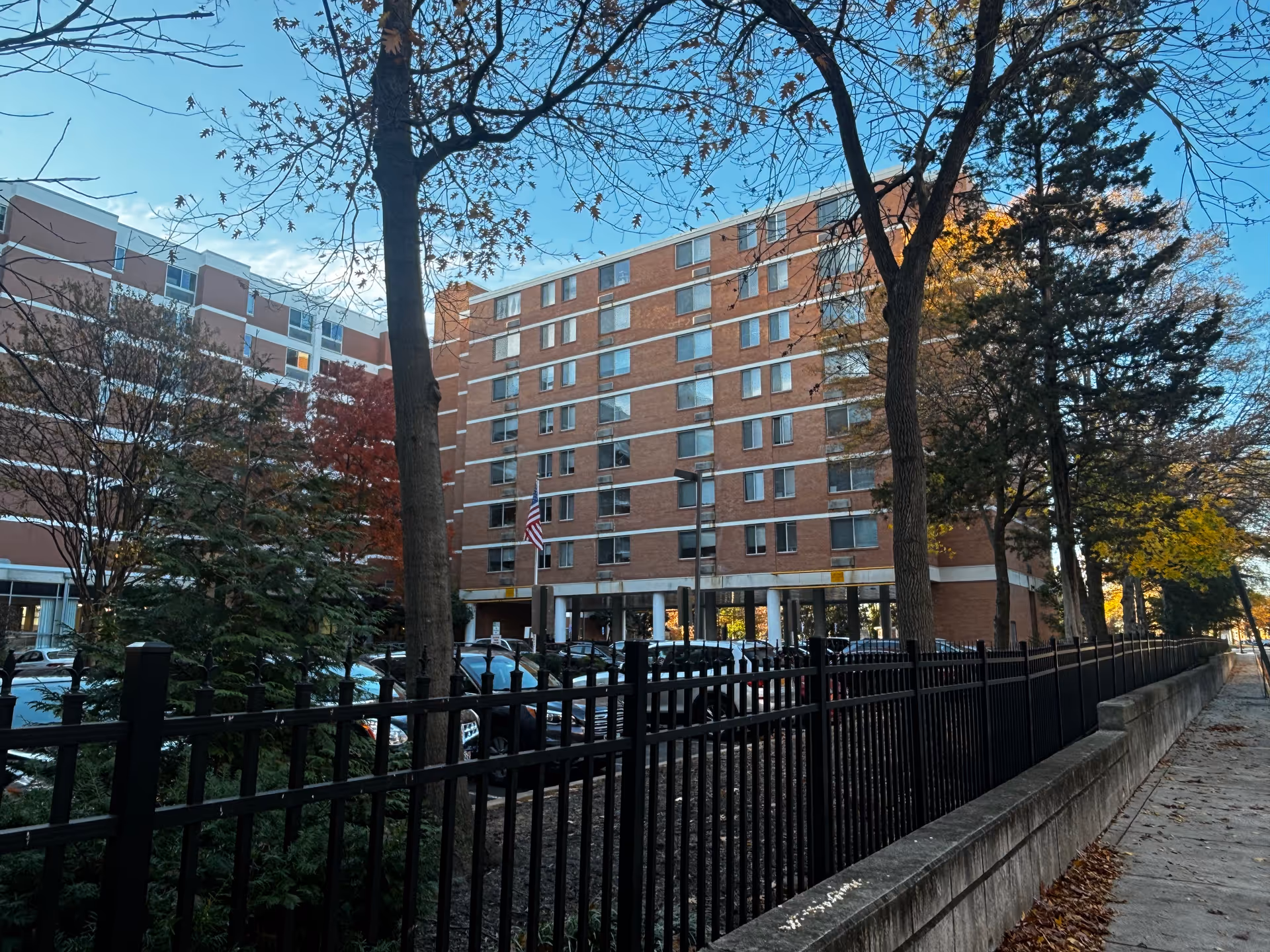 Exterior view of a multi-story brick building with several windows, surrounded by trees with autumn foliage. A black metal fence runs along the sidewalk in front of the building, and several parked cars are visible behind the fence. The sky is clear and blue.