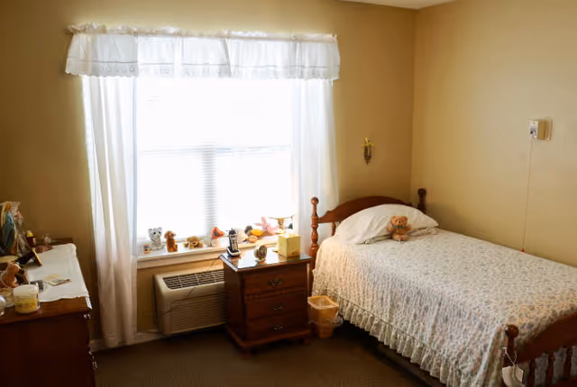 A small bedroom with a single wooden bed covered with a floral bedspread and a teddy bear on the pillow. Next to the bed is a wooden nightstand with a lamp, tissue box, and other small items. A window with white curtains lets in natural light, and a heating or air conditioning unit is below the window. There is a dresser with various personal items on the left side of the room. The walls are painted beige.
