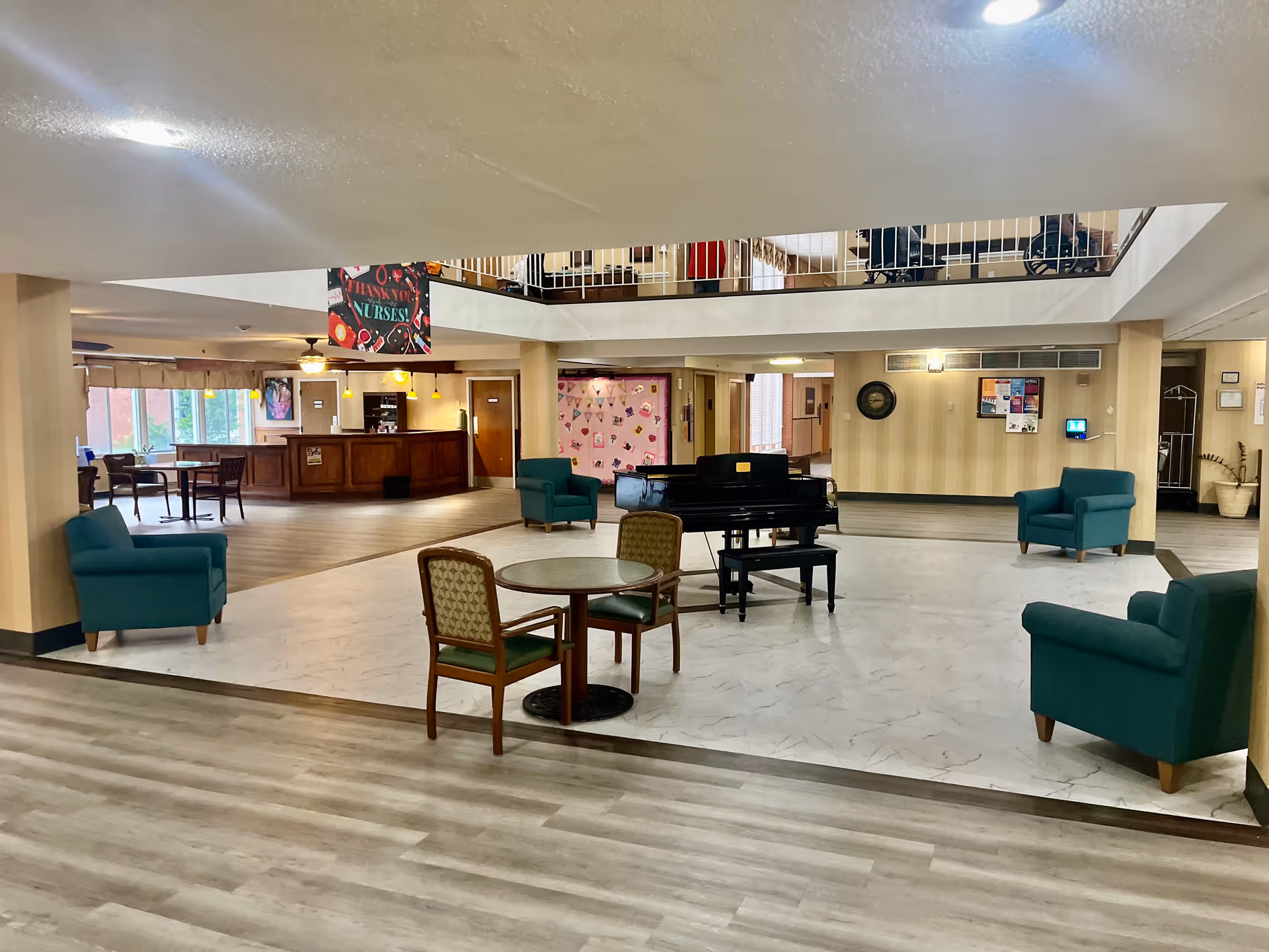 Open two-story nursing home lobby with seating, a grand piano, round tables and a reception desk.