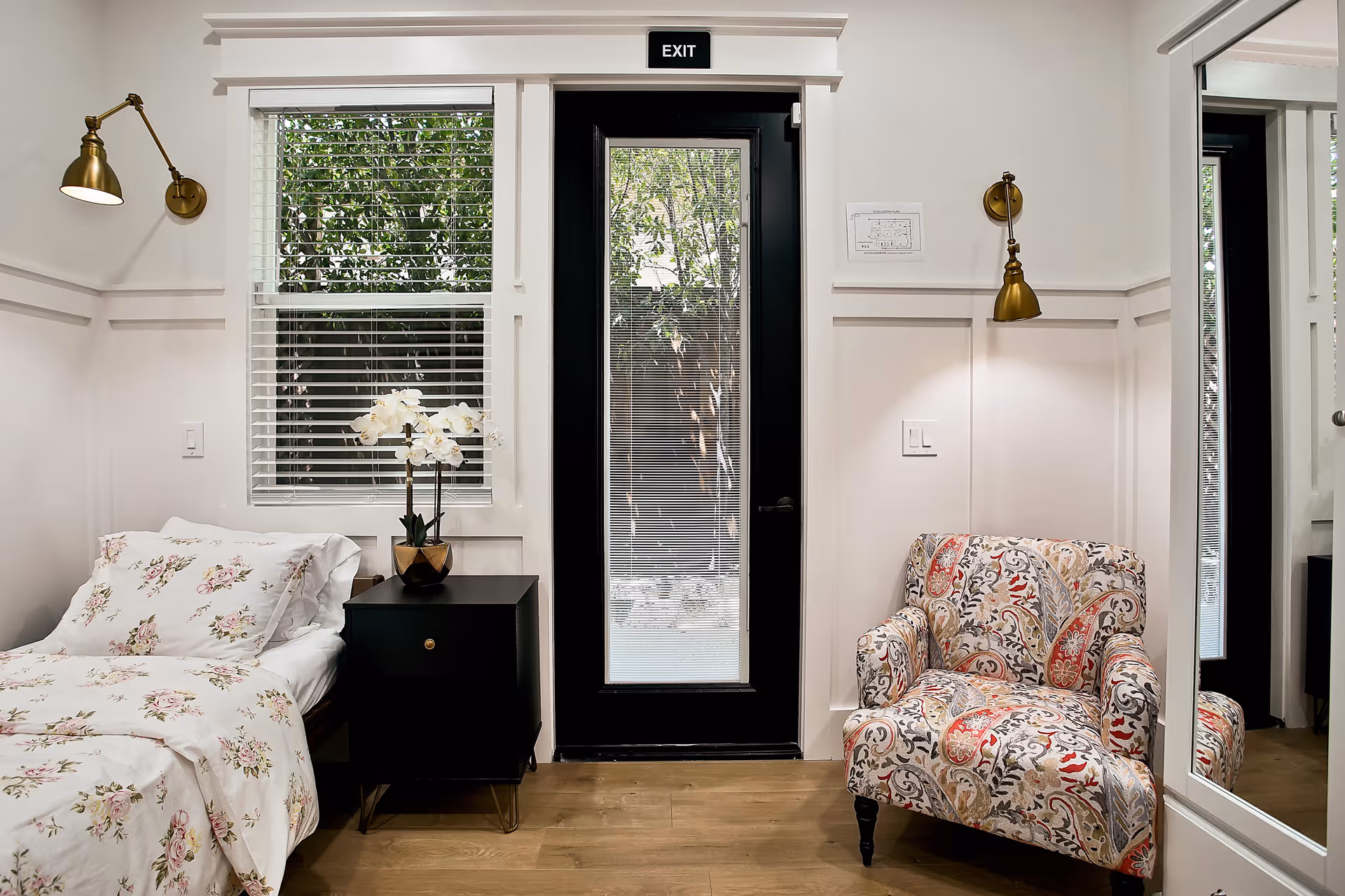 A cozy senior living bedroom with a floral-patterned bed on the left, a black nightstand with a white orchid plant, a window with white blinds, a black door with an exit sign above it, and a colorful patterned armchair on the right. The walls are white with decorative paneling and brass wall lamps above the bed and chair.