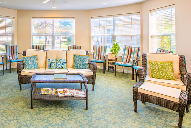 A bright and airy living room area with wicker furniture including a sofa and armchair with cream cushions and colorful patterned pillows. There is a wicker coffee table with magazines on the lower shelf. Several striped chairs are arranged along the windows, which let in natural light and offer a view of greenery outside. The room has a light blue patterned carpet and neutral-colored walls.