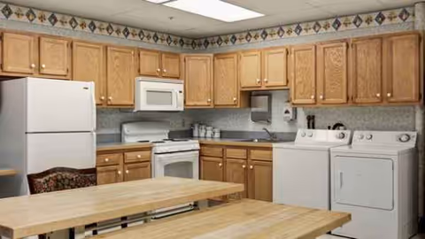 A well-lit senior living facility kitchen with wooden cabinets, appliances, and a wooden dining table.