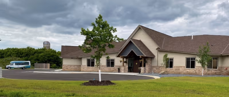 Single-story senior living building with a covered main entrance, a circular driveway, lawn and a parked shuttle under a cloudy sky.