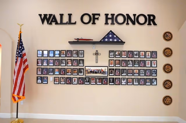 A wall display titled 'WALL OF HONOR' featuring framed photos of veterans arranged in rows, a folded American flag in a triangular case on a shelf, a cross, and a group photo in the center. To the left is a standing American flag, and to the right are five circular military insignias mounted vertically on the wall.