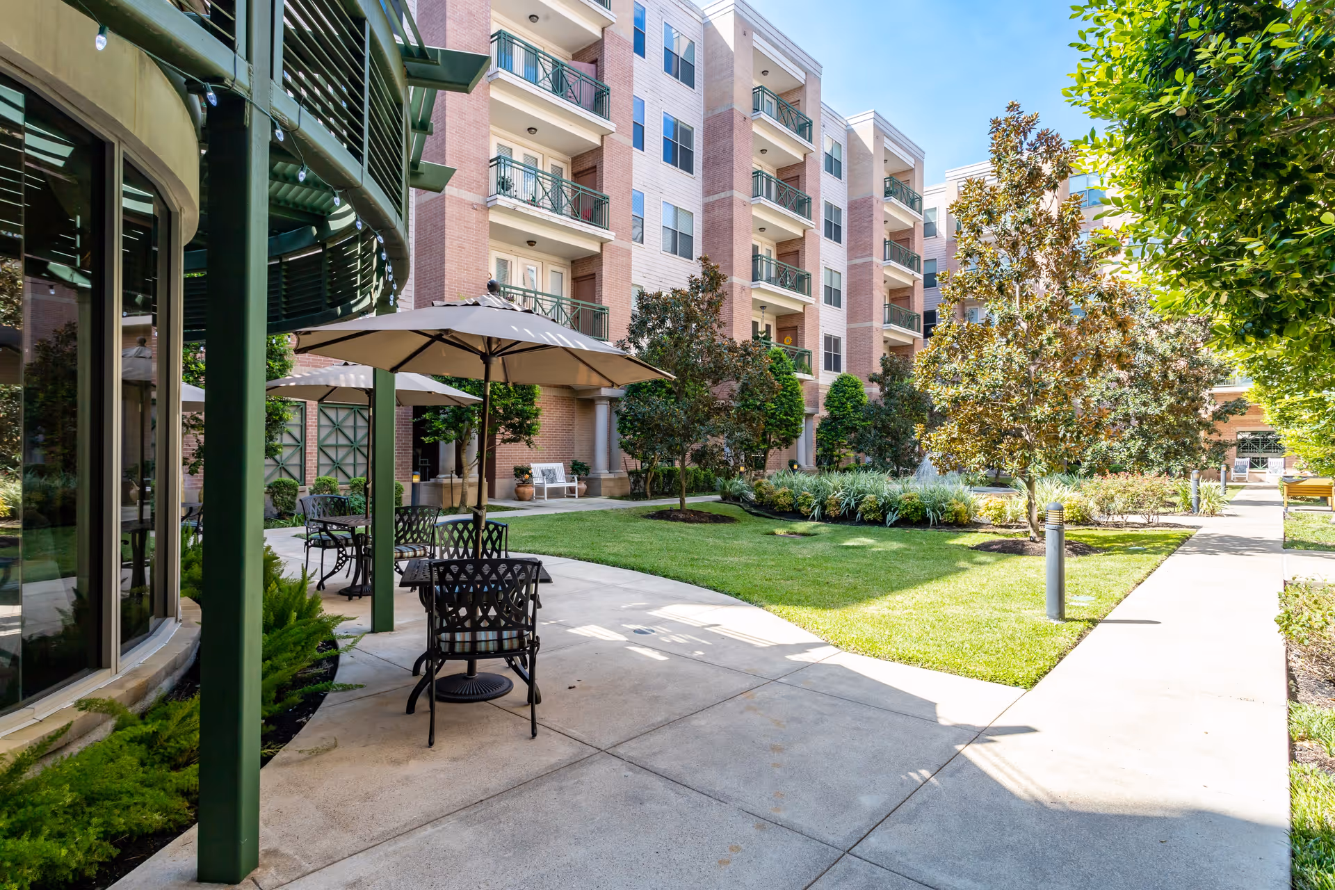 Outdoor courtyard area of The Village of Tanglewood featuring a paved walkway, green lawn, trees, and patio seating with tables and umbrellas next to a multi-story residential building.
