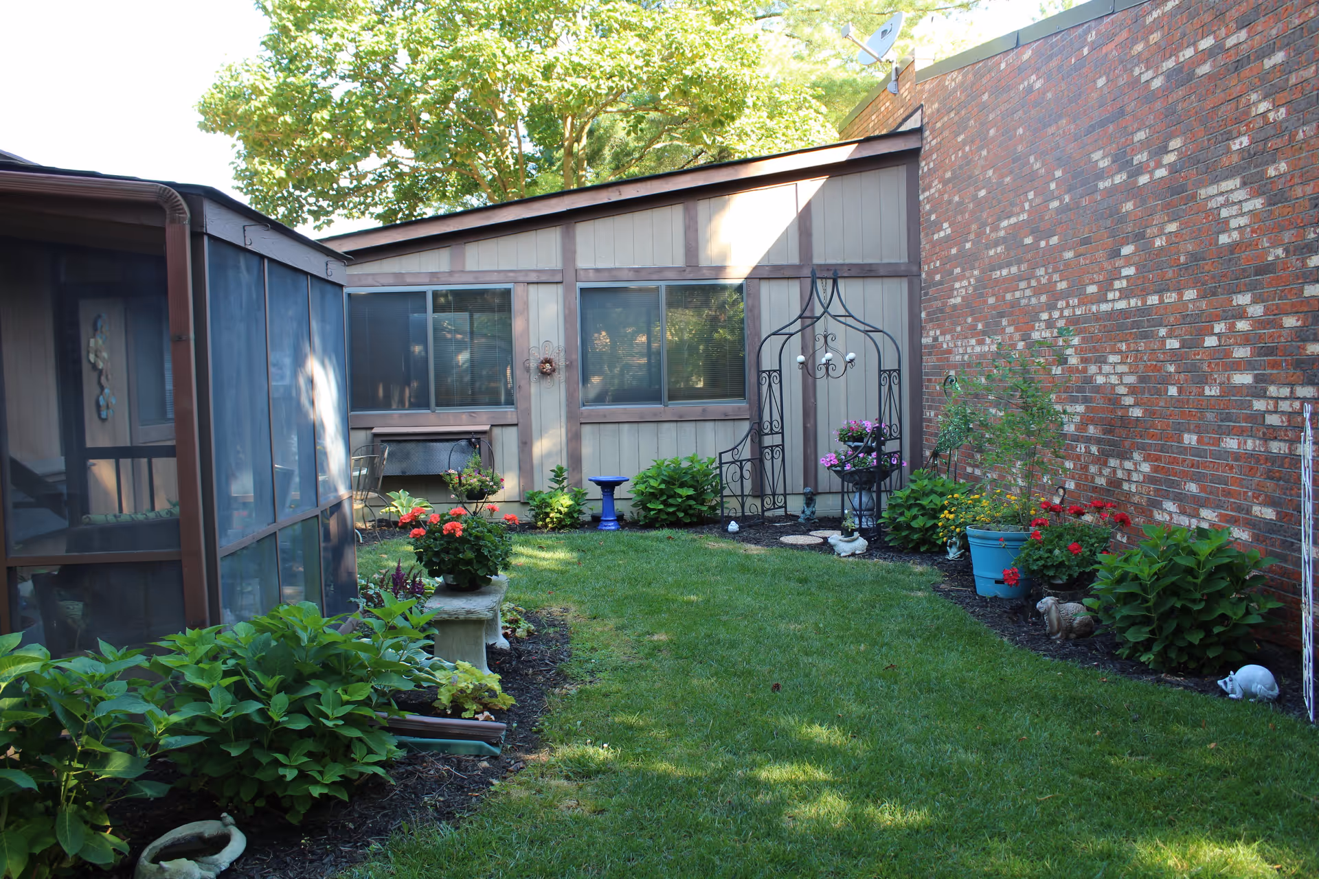 A small, well-maintained garden area with green grass, various flowering plants, and shrubs along the edges. There is a wooden building with two windows in the background, a brick wall on the right side, and a screened porch on the left. Decorative garden ornaments and a bench are also visible.