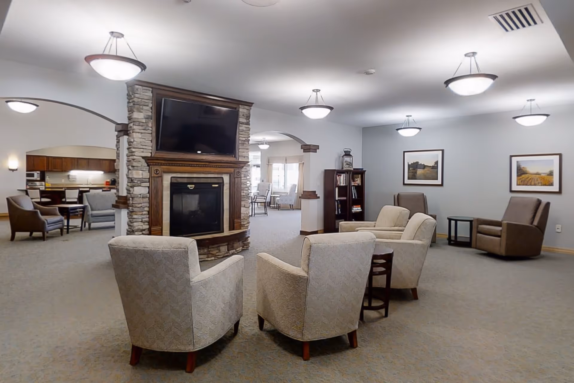 A spacious senior living facility common area with a stone fireplace and a mounted flat-screen TV. The room features several comfortable armchairs arranged around the fireplace, a bookshelf, framed landscape artwork on the walls, and soft overhead lighting. In the background, there is a dining area with tables and chairs visible through arched openings.