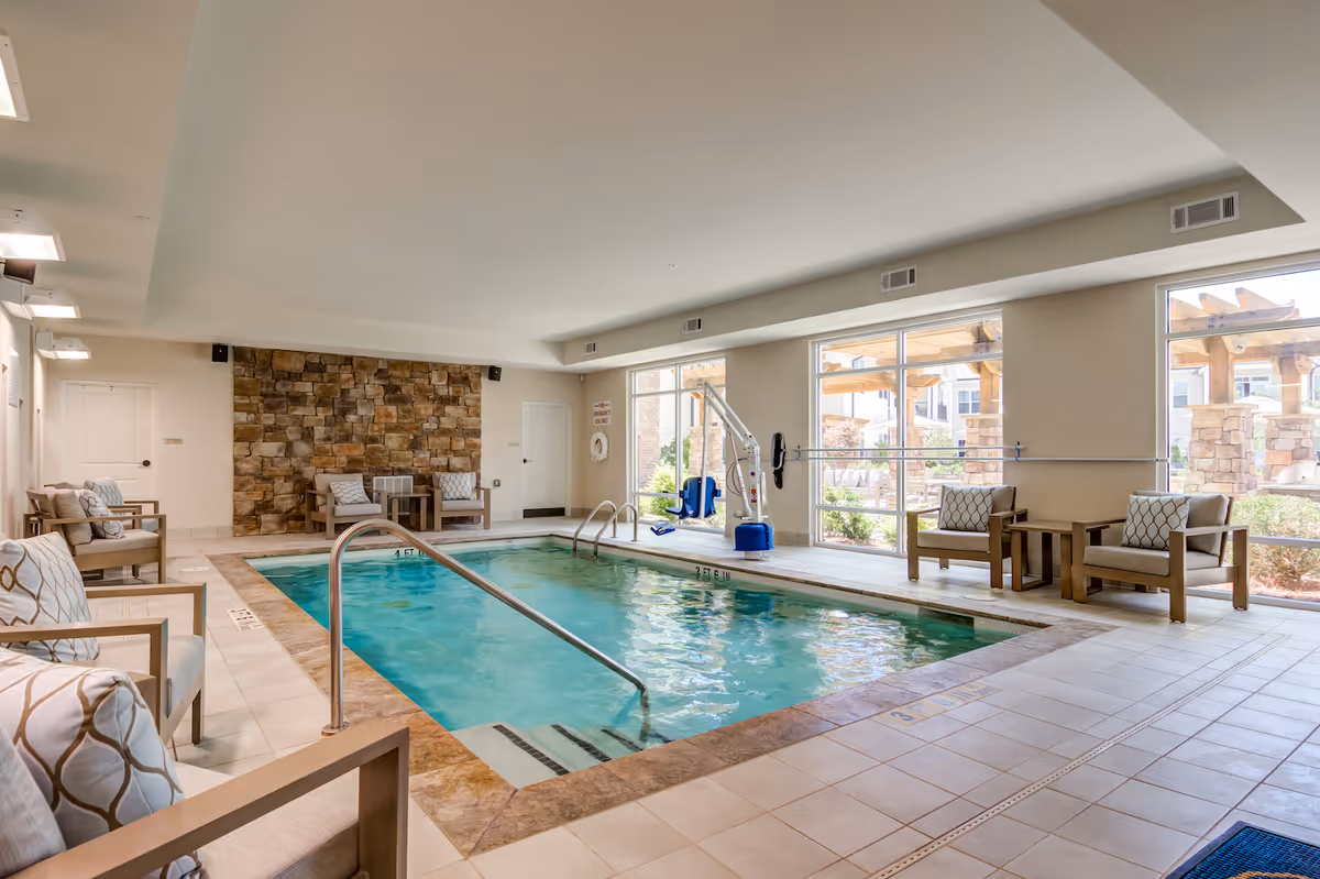 Indoor swimming pool area with clear blue water, surrounded by cushioned chairs and small tables. Large windows on one side let in natural light and show an outdoor pergola and garden area. The room has a stone accent wall and a pool lift for accessibility.