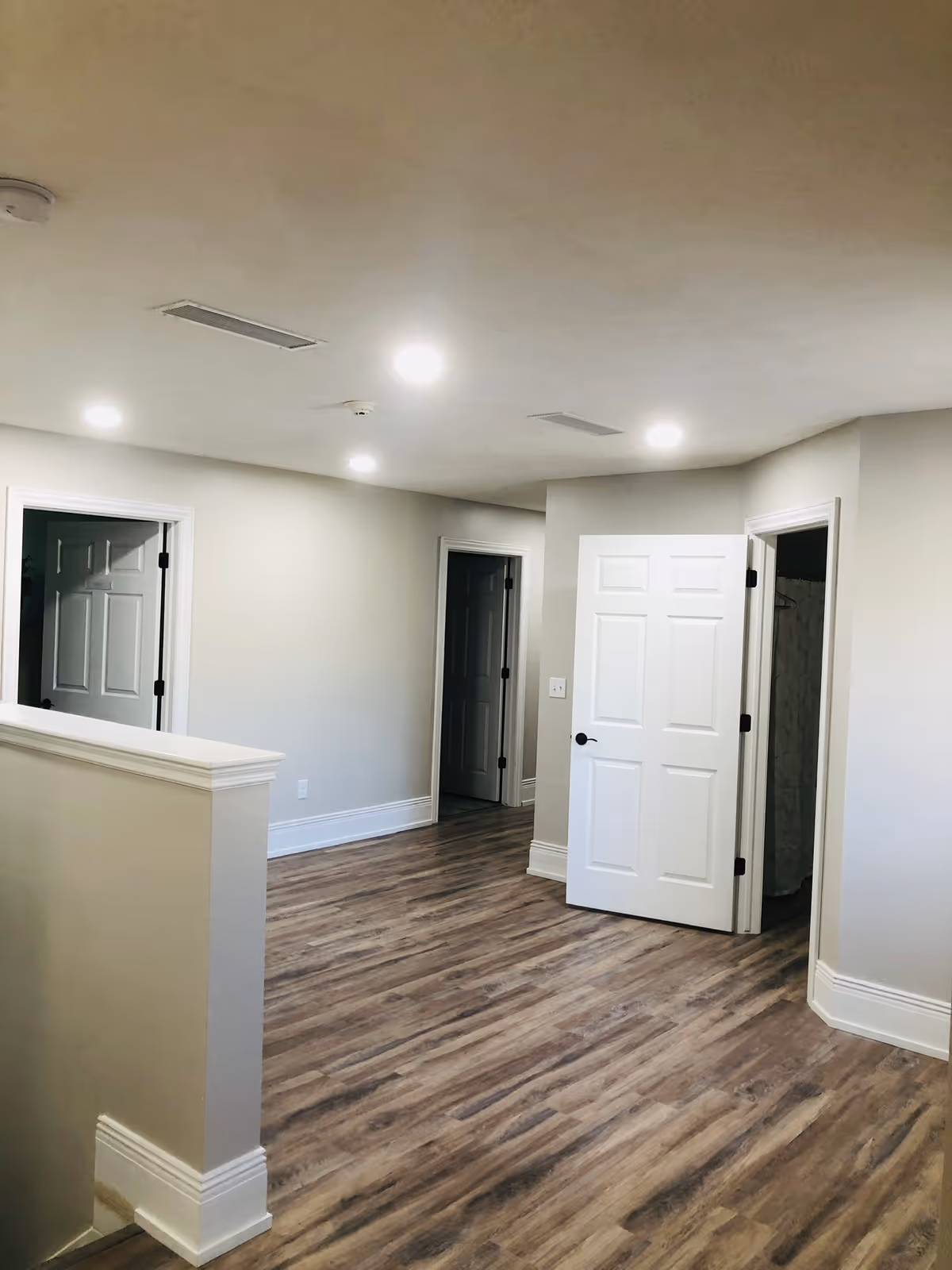 Interior hallway with wood flooring, beige walls, white trim, and multiple white doors, one of which is open revealing a shower curtain inside a bathroom.
