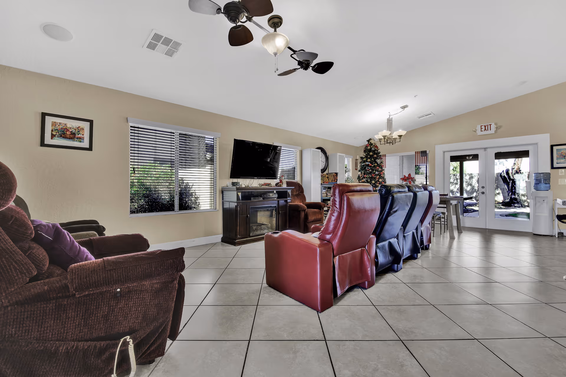 A spacious living room area in an assisted living facility featuring several recliner chairs arranged in rows facing a wall-mounted flat screen TV above a dark wooden cabinet. The room has tiled floors, beige walls, a ceiling fan with lights, and large windows with blinds. A decorated Christmas tree and an exit door with glass panels are visible in the background.