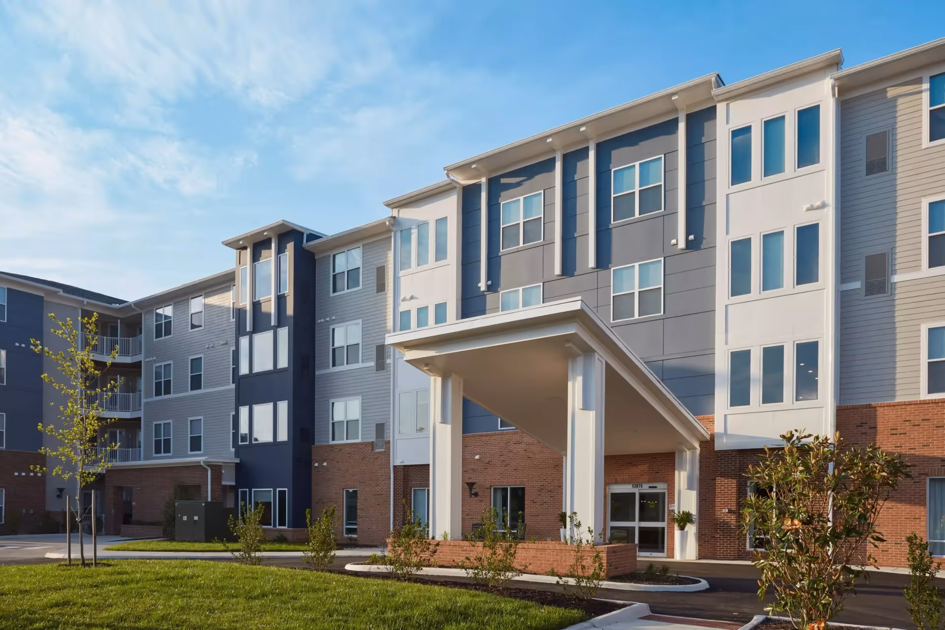 Exterior view of a modern multi-story senior living facility building with a covered entrance, large windows, and landscaped greenery under a blue sky.