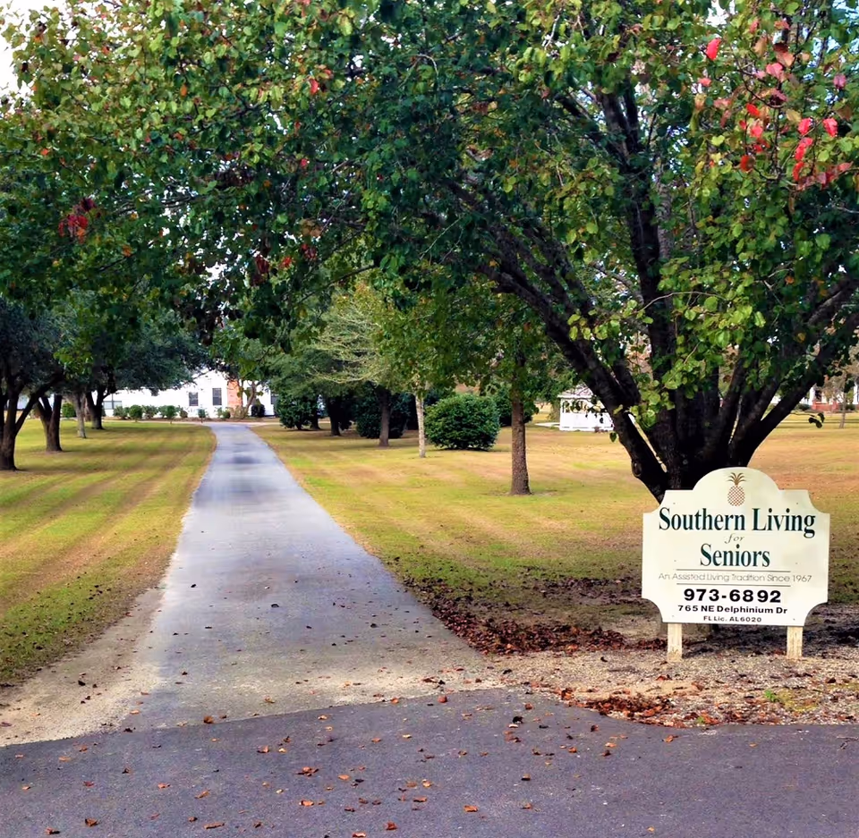 A paved driveway lined with trees on both sides leading to a building in the distance. On the right side of the driveway, there is a sign that reads 'Southern Living for Seniors, An Assisted Living Tradition Since 1967, 973-6892, 765 NE Delphinium Dr, FL Lic. AL 6020'. The area is grassy with some fallen leaves scattered on the driveway.