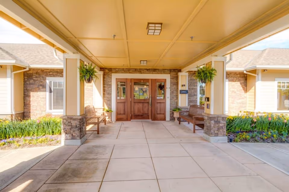 Covered entrance area of Arbor Oaks Terrace Memory Care Residence with double wooden doors, two wooden benches on either side, hanging plants, and flower beds along the sides of the building.