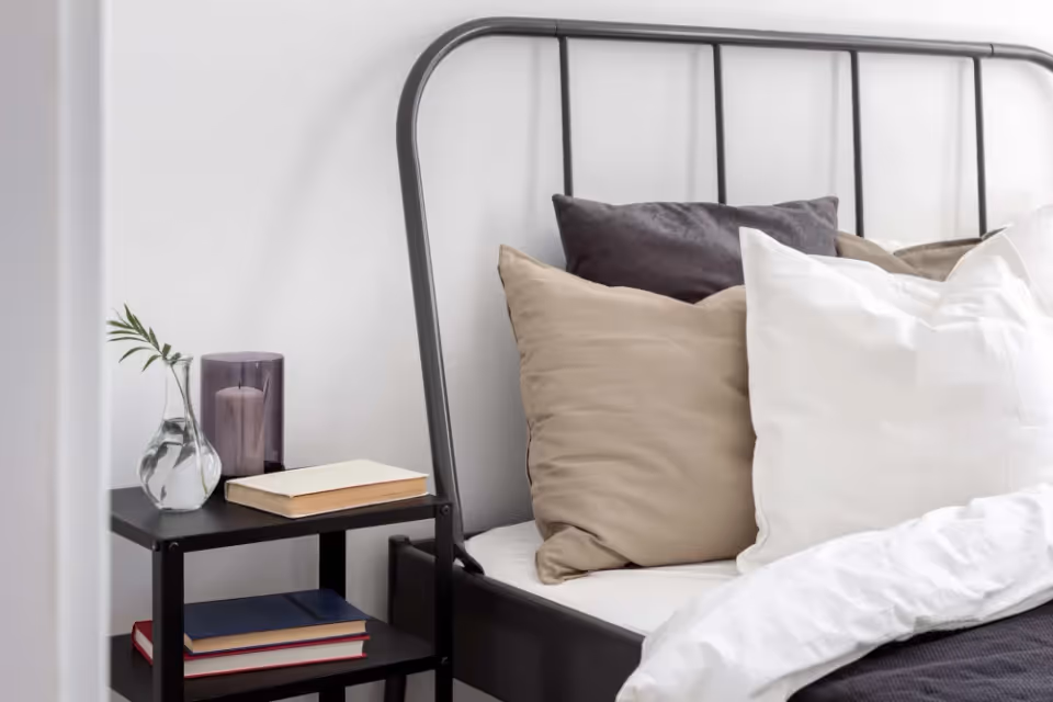 A tidy bedroom scene with a metal bed frame, white bedding, beige and white pillows, and a small black bedside table holding a vase, a candle, and books.