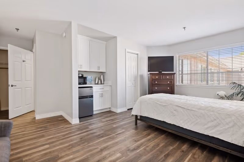 A bright bedroom with a large window covered by blinds, a bed with a white quilt, a wooden dresser with a flat-screen TV on top, and a small kitchenette area with white cabinets, a mini fridge, and a microwave. The room has wood flooring and white walls, with an open door leading to another room.