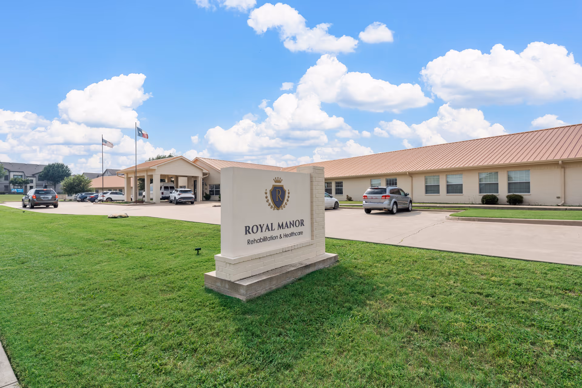 Exterior view of Royal Manor Rehabilitation and Healthcare building with a large sign in the foreground on a grassy lawn. The building has a beige exterior with a brown roof, several windows, and a covered entrance. There are a few cars parked in the driveway and two flagpoles with flags flying. The sky is blue with scattered clouds.