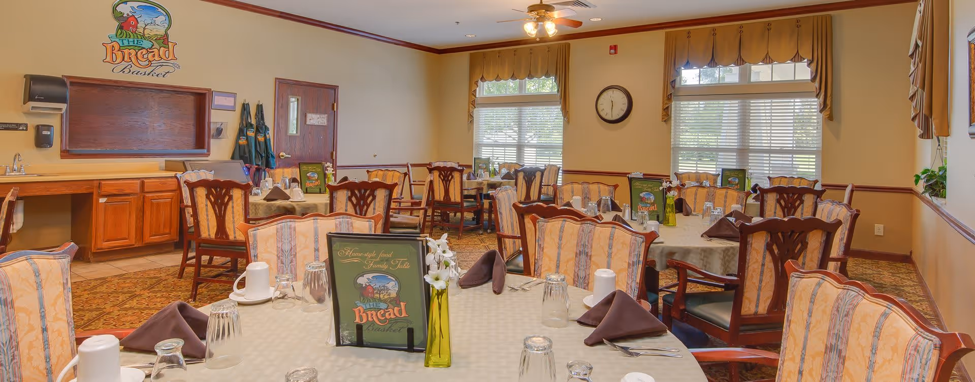 A dining room with multiple round tables covered with beige tablecloths, each set with cups, glasses, silverware, and brown folded napkins. The chairs have wooden frames with patterned upholstery. On the wall, there is a clock and windows with blinds and valances. A sign on the wall reads 'The Bread Basket' above a wooden counter with cabinets and a sink. The room has warm lighting and a carpeted floor.