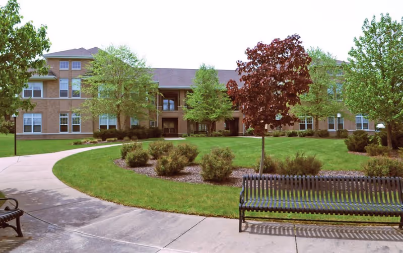 Outdoor view of a senior living facility with a curved concrete pathway, green lawn, small bushes, and several trees including one with red leaves. Two black metal benches are placed along the pathway. The building in the background is a two-story brick structure with multiple windows.