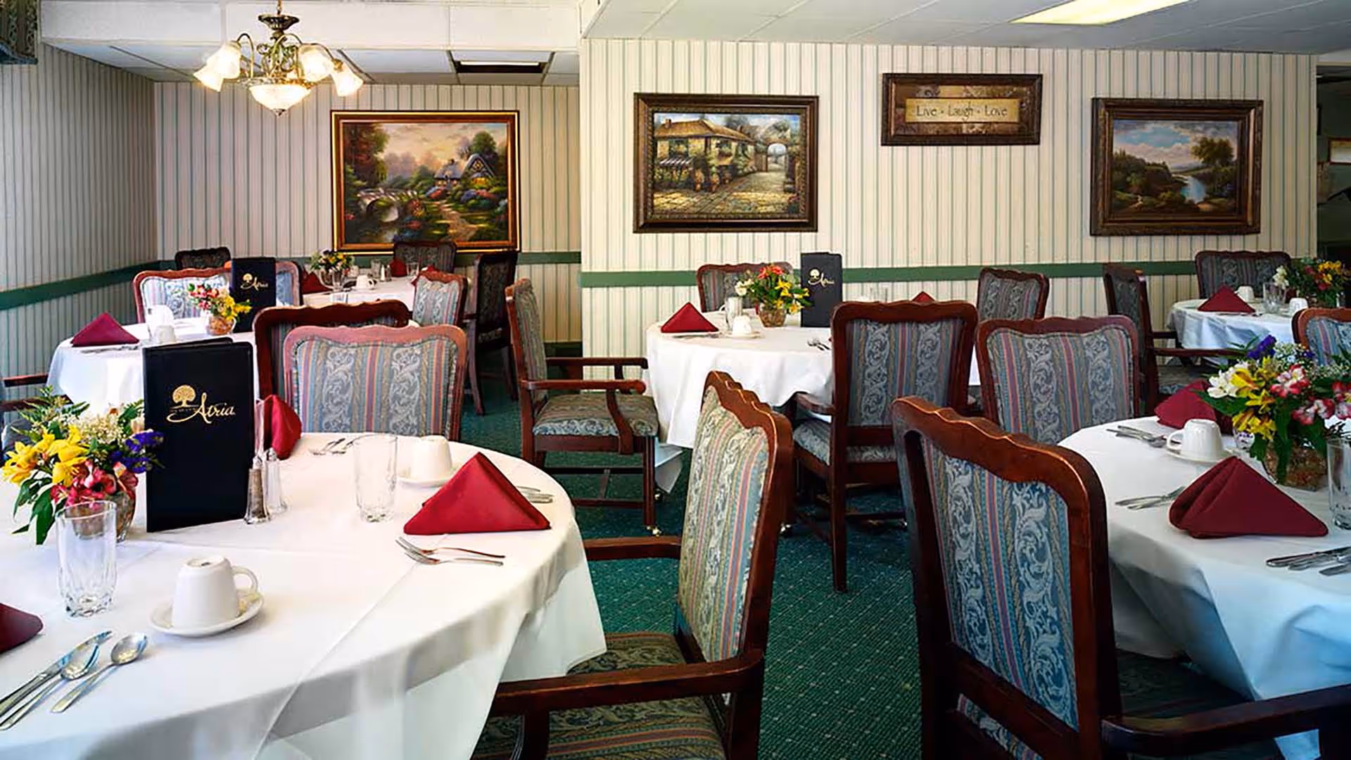 Dining room with round tables covered in white tablecloths, each set with red folded napkins, cups, glasses, and silverware. The chairs have patterned upholstery with wooden frames. There are flower arrangements on the tables and framed paintings on the striped wallpapered walls. A chandelier with multiple lights hangs from the ceiling.