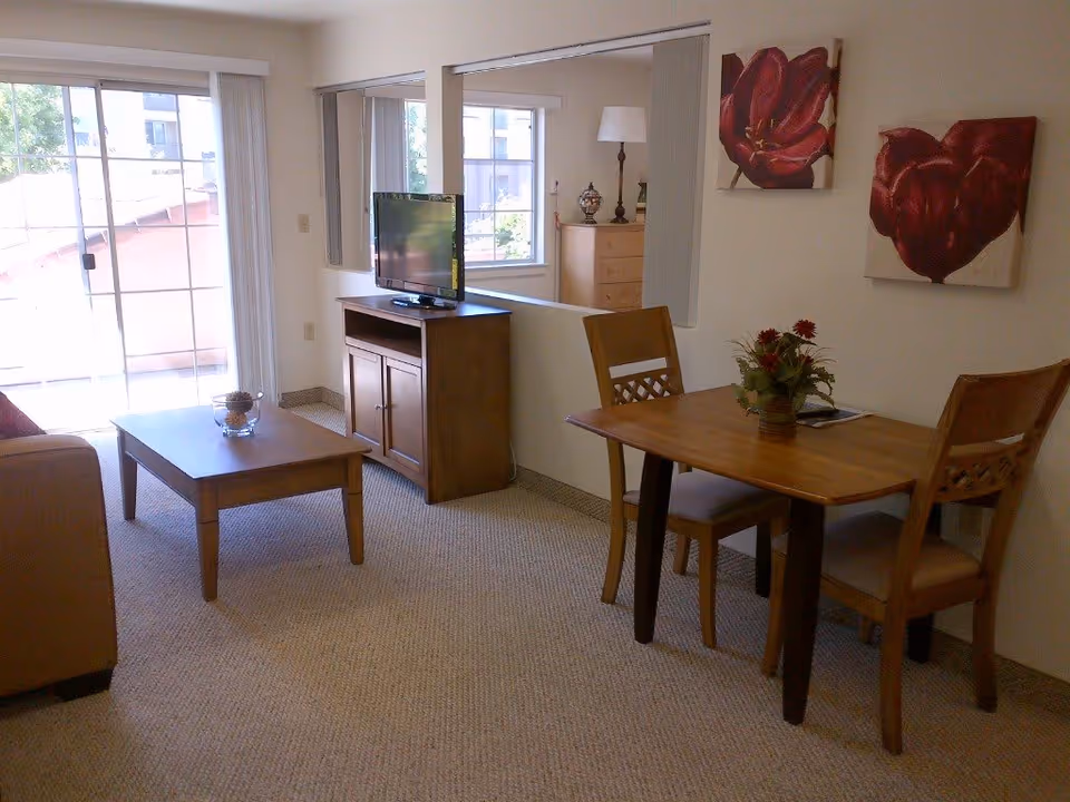 A cozy living and dining area in a senior living facility with a wooden coffee table, a TV on a wooden cabinet, a small dining table with two chairs, and two red floral paintings on the wall. There is a sliding glass door leading to an outdoor area and a window reflecting part of a bedroom.