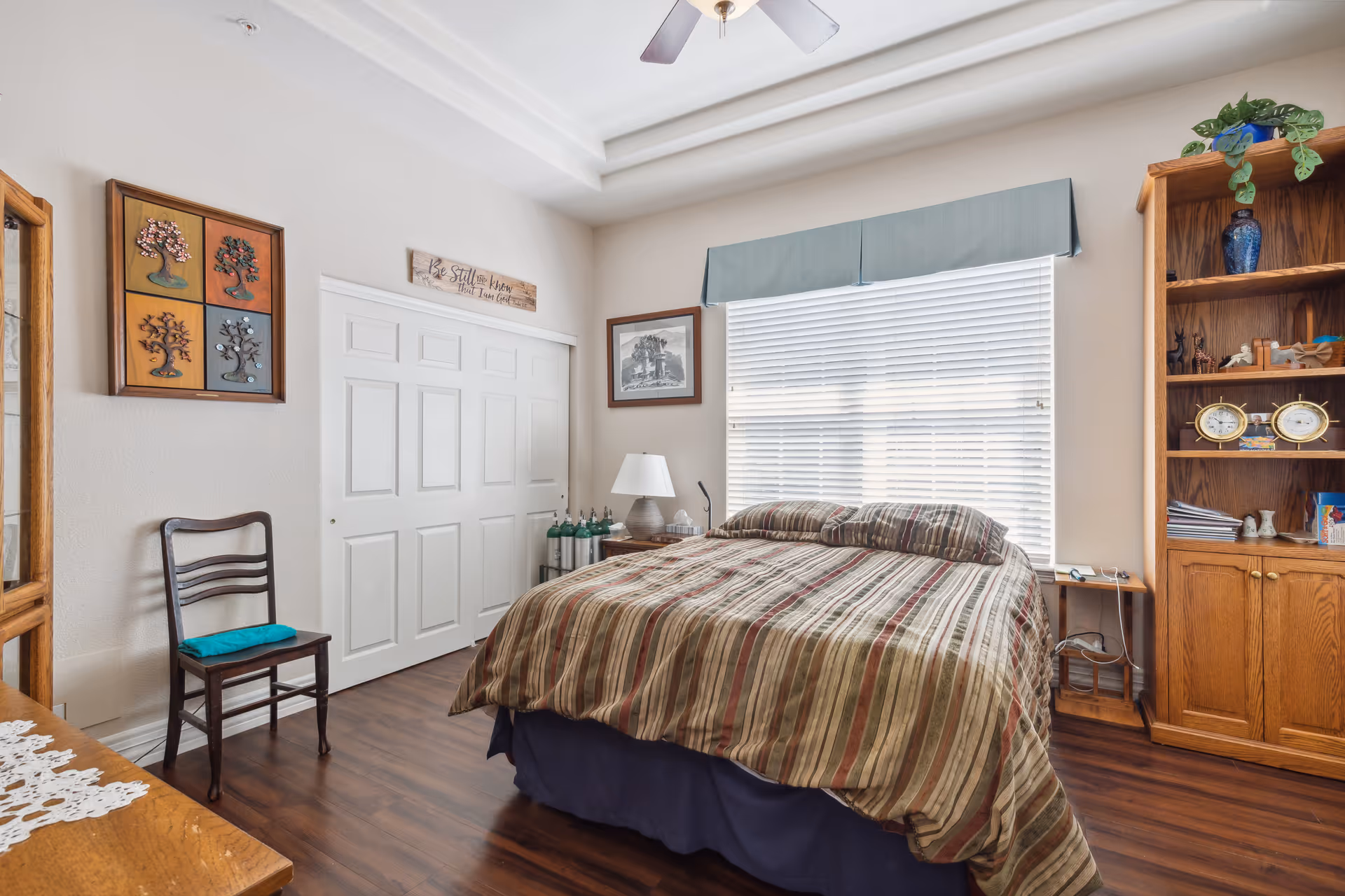 A bedroom with a bed covered in a striped comforter and two pillows, a wooden chair with a teal cushion, a wooden bookshelf with decorative items, a window with blinds and a blue valance, and a white closet door. The room has wood flooring and light-colored walls with framed artwork and a decorative sign above the closet.