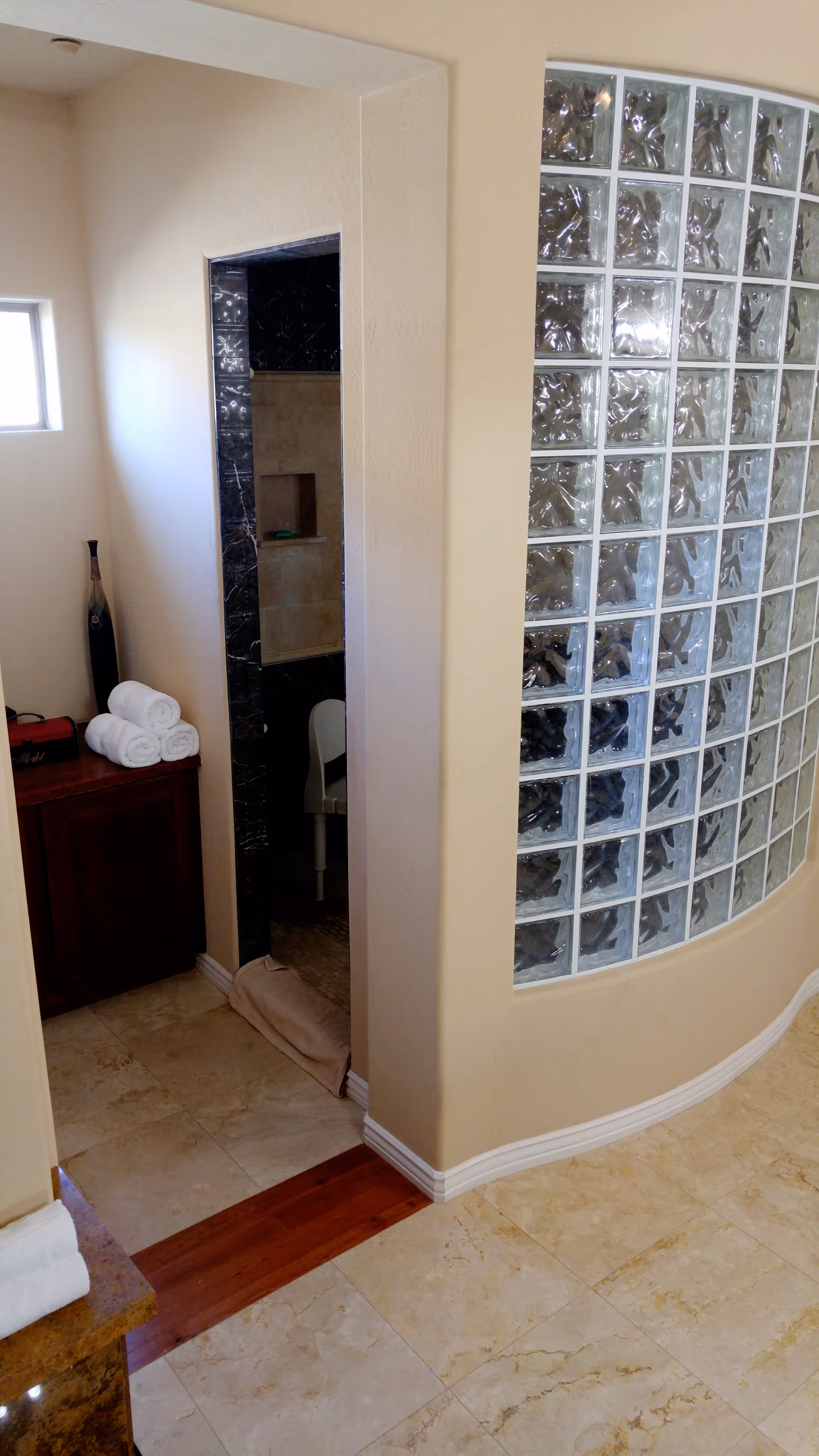 Curved glass-block wall beside a doorway into a tiled shower area with stacked towels on a wooden cabinet.