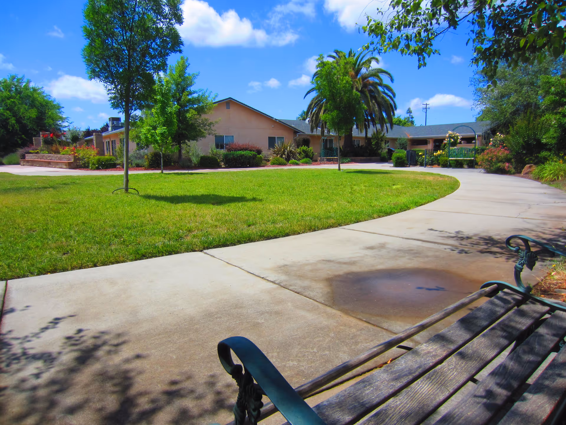 Outdoor view of a senior living facility with a curved concrete pathway, green lawn, trees, and a bench in the foreground. The building is visible in the background under a blue sky with scattered clouds.
