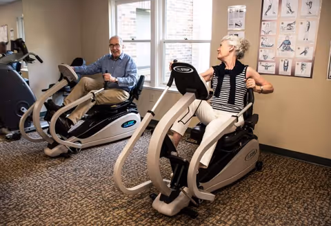 Two elderly individuals exercising on recumbent cross trainers in a well-lit fitness room with carpeted floor and exercise instruction posters on the wall.