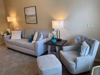 Well-lit sitting area with a sofa, armchair, ottoman, side tables, lamps, and wall art.