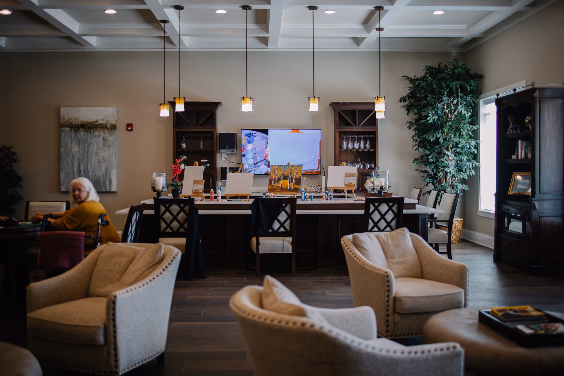 A cozy common area in an assisted living facility with several beige upholstered armchairs arranged around a central ottoman. In the background, there is a long table with art supplies and easels set up for painting activities. A large TV screen is mounted on the wall behind the table, flanked by two dark wooden cabinets. A large potted plant is near a window on the right side, and an elderly woman in a yellow sweater is seated at a table on the left side of the room. The room has a warm and inviting atmosphere with pendant lights hanging from a coffered ceiling.