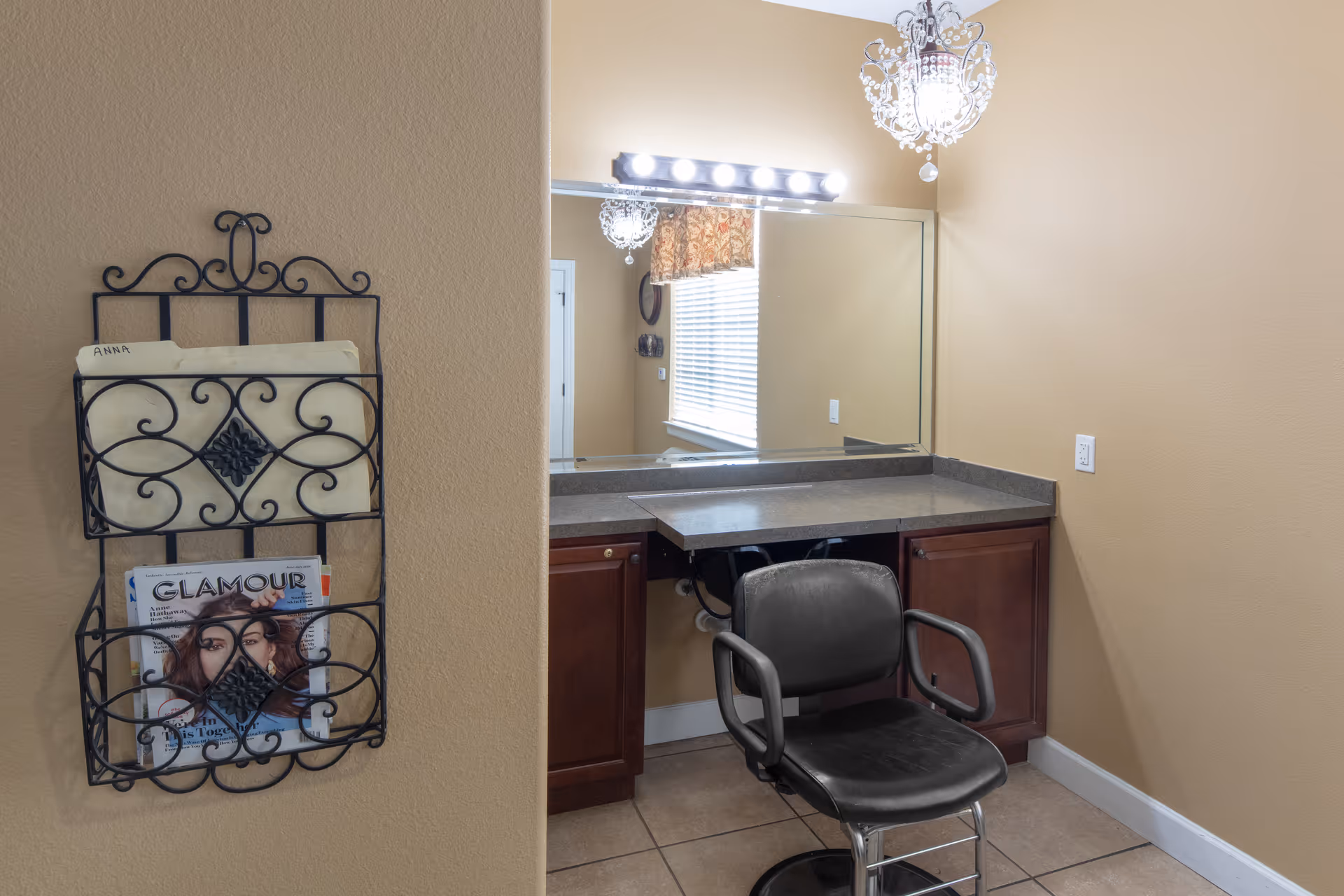 A small vanity area with a large mirror, overhead lights, and a black salon chair in front of a countertop with wooden cabinets underneath. On the left wall, there is a decorative black metal magazine rack holding some papers and a Glamour magazine. The walls are painted beige and the floor is tiled.