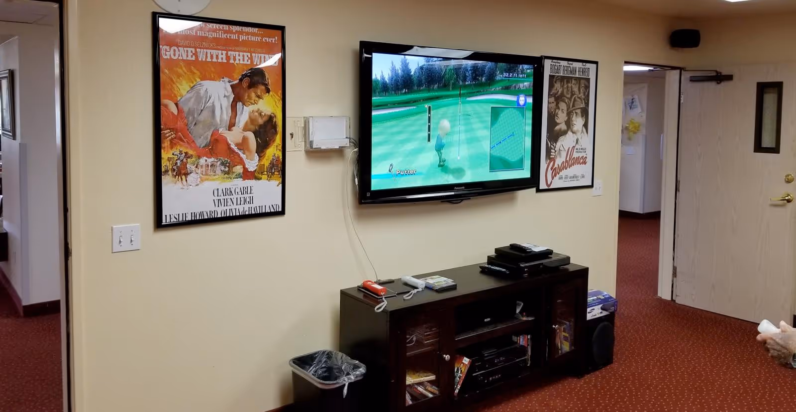 A living room area in a senior living facility with a wall-mounted flat-screen TV displaying a golf video game. Below the TV is a dark wooden entertainment cabinet holding various electronic devices and game cases. On the wall, there are two framed classic movie posters: Gone With The Wind and Casablanca. The room has beige walls and red carpet with a dotted pattern. A trash can is placed near the cabinet, and a person holding a game controller is partially visible on the right side.