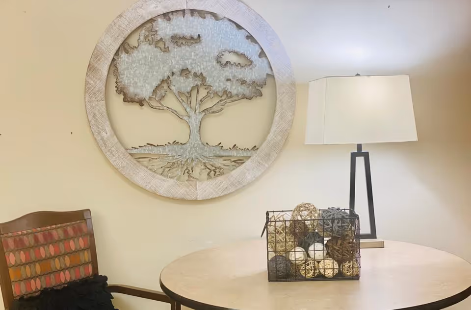 A cozy corner with a round wooden table holding a decorative wire basket filled with various textured balls, a modern table lamp with a rectangular shade, and a wooden chair with a patterned cushion. On the wall above the table is a large round wall art piece featuring a tree design.