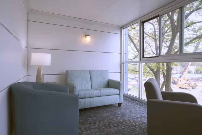 A small seating area with a light blue loveseat, two gray armchairs, a side table with a lamp, and a large window showing trees and construction equipment outside.