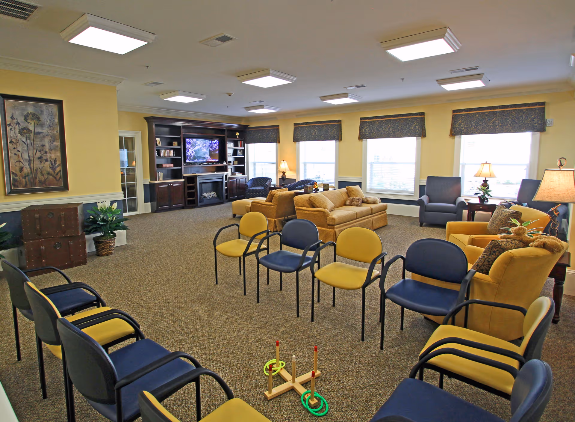 A spacious senior living facility common area with a circle of yellow and blue chairs arranged around a ring toss game. The room features beige carpeting, yellow walls, multiple windows with valances, a large dark wood entertainment center with a TV, and several comfortable sofas and armchairs. There are also lamps and decorative plants placed around the room.