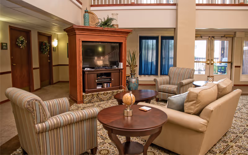 A cozy living room area in a senior living facility with a beige leather sofa, two striped armchairs, a wooden TV cabinet with a flat-screen television, two round wooden coffee tables, decorative vases, and blue curtains on the windows. The room has patterned carpet flooring and doors leading to other rooms and an outdoor area.