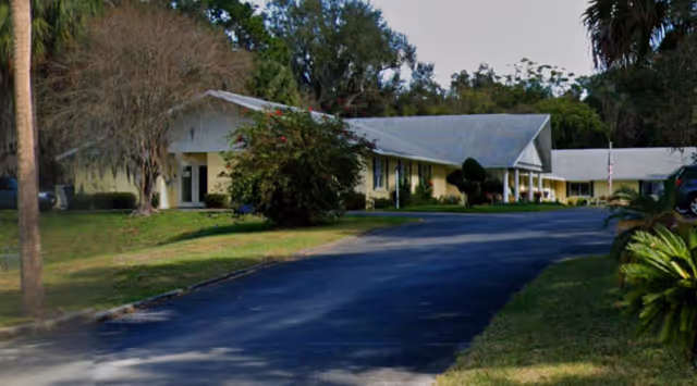 A single-story yellow building with a gray roof surrounded by trees and greenery, with a paved driveway leading up to it under a clear sky.
