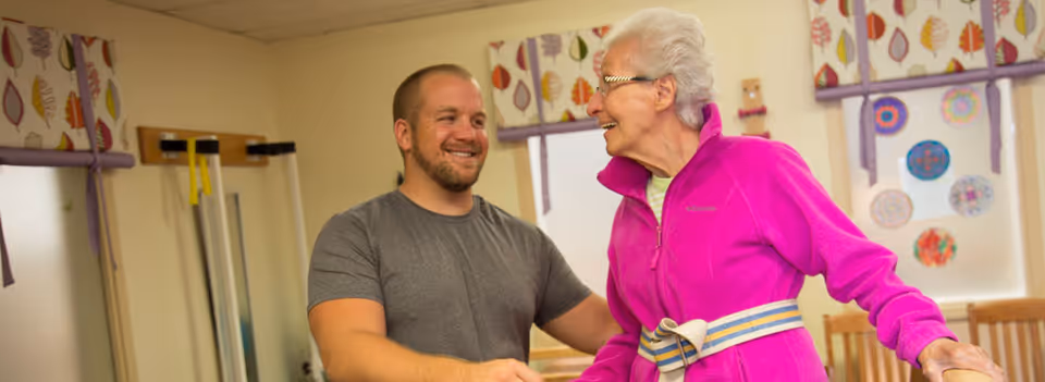 An elderly woman wearing a bright pink jacket and a physical therapy gait belt is smiling and interacting with a male caregiver in a room with light-colored walls and windows with patterned curtains.