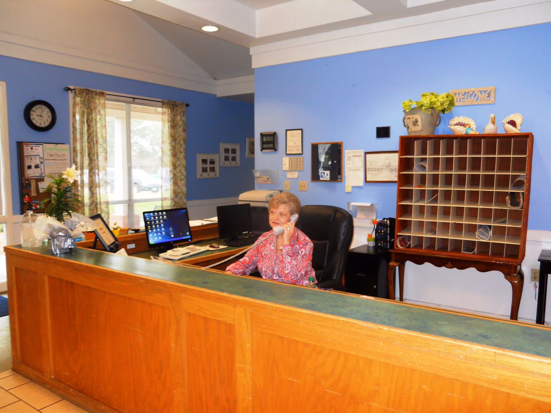 Reception area of an assisted living facility with a woman sitting behind a wooden desk talking on the phone. The desk has a computer, keyboard, and some paperwork. Behind her is a blue wall with a wooden mail organizer, a welcome sign, and various framed documents. A window with floral curtains is visible to the left.
