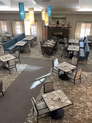 A dining room with multiple tables set with white cups, plates, and silverware. The room features blue cushioned bench seating along the walls, individual chairs around the tables, and modern hanging pendant lights in blue and yellow. There is a fireplace decorated with holiday garland and figurines, and windows with sheer curtains letting in natural light.